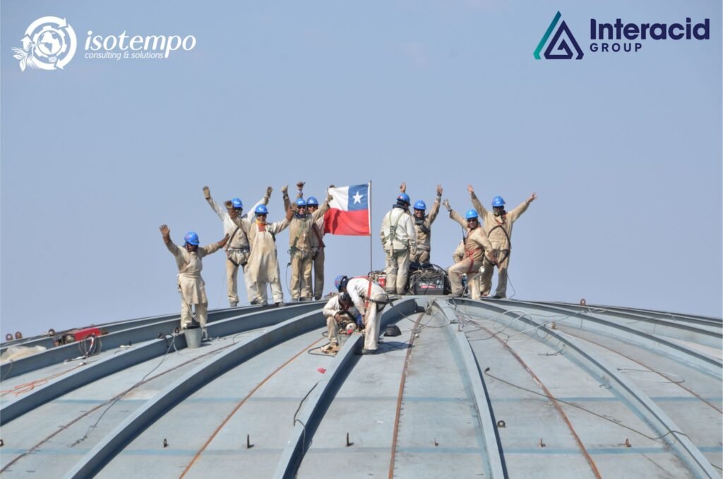 Trabajadores industriales con cascos azules celebrando sobre estructura metálica junto a la bandera de Chile – Isotempo Consulting & Solutions e Interacid Group